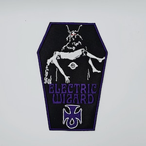 May include: Embroidered patch in a coffin shape, featuring the words "ELECTRIC WIZARD" in purple. The black patch depicts a figure holding a person, with white and purple detailing.