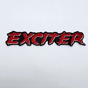 May include: Embroidered patch featuring the word "EXCITER" in red, with a white outline, set against a black backdrop. The patch has a distressed, jagged design, suitable for clothing or accessories.