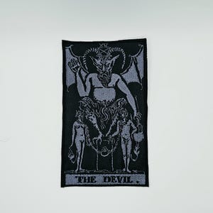 May include: A rectangular embroidered patch featuring a black and light grey design of The Devil tarot card. The image depicts a horned figure with wings, a pentagram above its head, and two chained figures below, with the text THE DEVIL at the bottom.