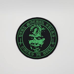 May include: Round black patch with green text and a green skull design. The text around the edge reads "YOUR MOTHER SUCKS" and "COOKS IN HELL". Two green crosses are on either side of the skull.