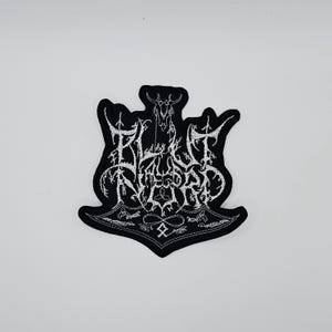 May include: Black and white embroidered patch with the text "BLEEDING NORTH" in a stylized font. The patch features a horned skull and other decorative elements. The patch is shaped like a hammer.