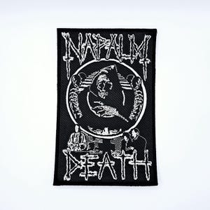 May include: Black and white embroidered patch featuring the word "NAPALM" above a circular design with a hooded figure and skeletons. The word "DEATH" is below the circle. The patch is rectangular.