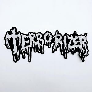 May include: A black and white embroidered patch with the word "TERRORIZER" in a dripping, stylized font. The patch has a black border and is set against a white background.