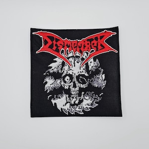 Puede incluir: Parche cuadrado negro con un diseño de calavera blanca y un logotipo rojo "Dismember". La calavera está rodeada por un diseño blanco y dentado. Este parche es probablemente para ropa o accesorios.