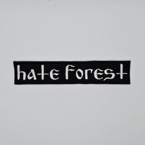 Pode incluir: Um patch retangular preto com as palavras "hate Forest" bordadas em branco, com letras estilizadas. O patch está centrado em um fundo branco. O texto está em uma fonte gótica.