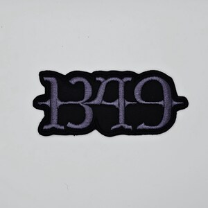 Peut inclure: Écusson brodé noir avec le nombre "1349" en police gothique. Les chiffres sont de couleur violet clair et cousus sur un fond noir. L'écusson a une forme stylisée unique.