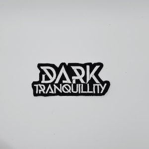 Può includere: Una toppa ricamata in bianco e nero con le parole "DARK TRANQUILLITY". Il testo è in un font stilizzato, con la parola "DARK" sopra "TRANQUILLITY". La toppa ha un bordo nero ed è posta su uno sfondo bianco.