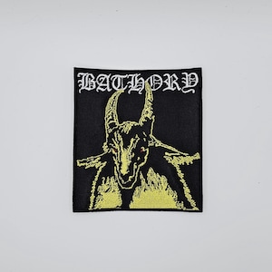 May include: Black square patch with the word "BATHORY" in white gothic lettering above a yellow illustration of a horned creature with red eyes. The patch is likely for clothing or accessories.