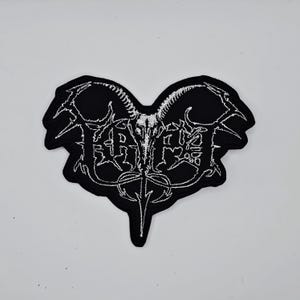 May include: Black and white embroidered patch with a heart-shaped design. The patch features a stylized goat skull and the word "FAITH" in a gothic font. The patch is likely for clothing or accessories.