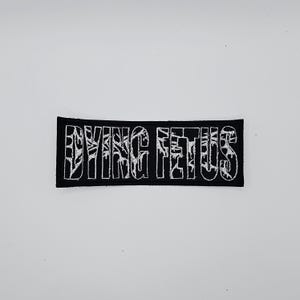 Könnte beinhalten: Schwarzer rechteckiger Aufnäher mit dem weißen gestickten Text "DYING FETUS". Der Text hat ein stilisiertes, strukturiertes Design. Der Aufnäher ist wahrscheinlich für Kleidung oder Accessoires bestimmt.