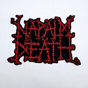 May include: Black patch with the words "NAPALM DEATH" in red, stylized, jagged lettering. The patch has a unique, irregular shape, and the text appears to be embroidered or stitched onto the black fabric. The background is plain white.