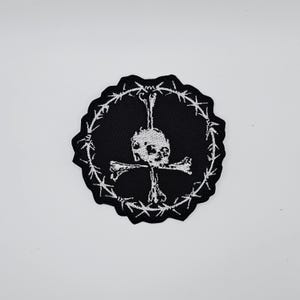 Op de afbeelding: Zwarte patch met een wit ontwerp van een schedel en gekruiste botten. De schedel is gecentreerd, met een bot dat omhoog steekt en twee botten die een kruis vormen. Het ontwerp is omcirkeld door een witte prikkeldraadrand.