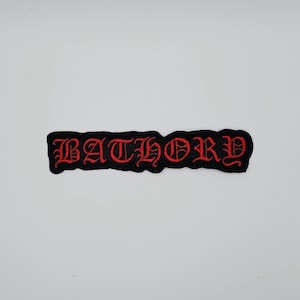 May include: Black embroidered patch featuring the word "BATHORY" in red gothic script. The patch has a black border and a rectangular shape. The text is in a bold, stylized font.