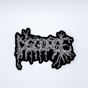 May include: Black embroidered patch with the word "CORPSE" in a stylized, gothic font. The white thread creates a textured, intricate design against the black fabric. The patch has a unique, irregular shape.