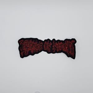 May include: Black embroidered patch with the text "CRADLE OF FILTH" in red. The patch has a jagged edge and is rectangular in shape. The text is in a gothic font.