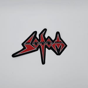 Puede incluir: Parche bordado con la palabra "SODOM" en una fuente estilizada y angular. El texto es rojo con detalles blancos y un borde negro. El parche tiene un diseño simétrico, adecuado para aplicar en ropa o accesorios.