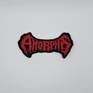 Può includere: Una toppa ricamata nera e rossa con la scritta "AMORPHIS" in un carattere gotico stilizzato. Il testo è rosso, con un bordo nero. La toppa ha una forma curva distintiva.
