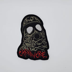 Op de afbeelding: Zwarte patch met een gasmasker ontwerp, witte ooglenzen en een centraal filter. Het woord "CARNIVORE" is in het rood geborduurd. De patch is waarschijnlijk bedoeld voor kleding of accessoires.