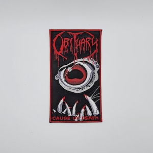 Può includere: Toppa ricamata con la scritta "Obituary" in rosso gocciolante. Sotto, una grafica dettagliata di un occhio con iride rossa e denti affilati. Il testo "CAUSE OF DEATH" è in basso, il tutto su sfondo nero.
