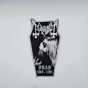 Könnte beinhalten: Schwarz-weißer Aufnäher mit dem Bandnamen "Mayhem" in gotischer Schrift, über einem Bild eines Gesichts. Der Aufnäher ist sargförmig mit Fledermausflügel-Akzenten und dem Wort "DEAD" darunter, mit den Jahren 1969-1991.