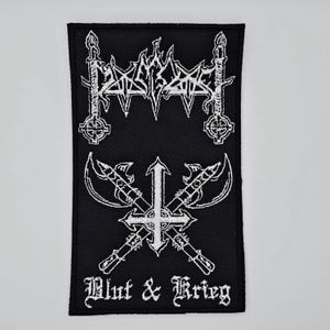 May include: Black rectangular patch with white embroidered designs. The top features a stylized logo with two candle-like shapes. The bottom has crossed axes with a cross and the words "Blut & Krieg".