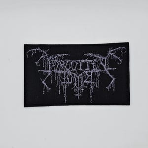 May include: Black rectangular patch with the text "FORGOTTEN TOMB" in a stylized, gothic font. The text is in a light grey color, and the patch has a black border.