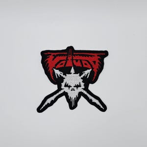 May include: Embroidered patch with a striking design in red, white, and black. The design features a red, wing-like shape at the top, a white skull-like figure in the centre, and two white, sword-like shapes extending downwards.
