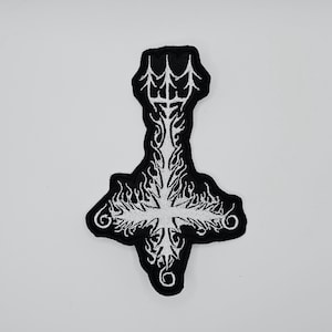 May include: Black and white embroidered patch featuring an inverted cross design. The patch has a black border and a detailed white cross with flame-like accents. The patch is likely for clothing or accessories.