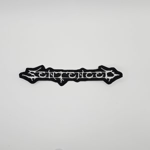 Op de afbeelding: Een zwart-wit geborduurde patch met het woord "SENTENCED" in een gotisch lettertype. De patch heeft een pijlvorm met puntige uiteinden. De tekst is wit en de patch staat op een witte achtergrond.