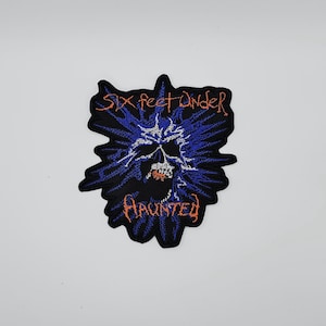 May include: Embroidered patch featuring a skull design with the words "SIX FEET UNDER" and "HAUNTED" in orange lettering. The skull is white with red accents, set against a dark blue and black background.