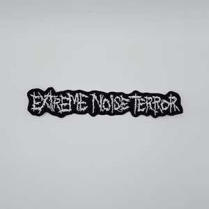 May include: A black embroidered patch featuring the text "EXTREME NOISE TERROR" in white, jagged lettering. The patch has a black border and a rectangular shape. It is designed to be sewn onto clothing or bags.
