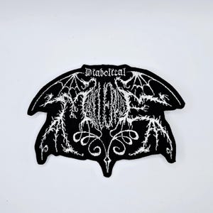 Könnte beinhalten: Schwarzer, gestickter Aufnäher mit dem Wort "Diabolical" in gotischer Schrift. Der Aufnäher zeigt ein fledermausartiges Design mit komplizierten weißen Details, einschließlich Flügeln und einer zentralen, stilisierten Illustration. Der Aufnäher hat eine einzigartige Form.