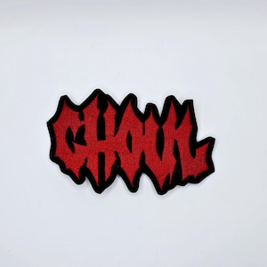Może przedstawiać: Czerwona, haftowana naszywka z napisem "GHOUL" w stylizowanej, poszarpanej czcionce. Tekst jest obrysowany na czarno. Naszywka jest przeznaczona do odzieży lub akcesoriów.
