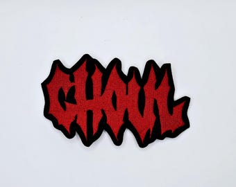 Może przedstawiać: Czerwona, haftowana naszywka z napisem "GHOUL" w stylizowanej, poszarpanej czcionce. Tekst jest obrysowany na czarno. Naszywka jest przeznaczona do odzieży lub akcesoriów.