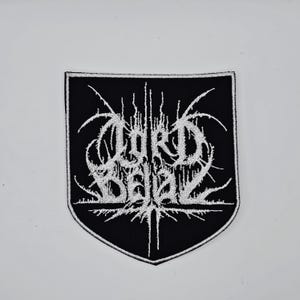 May include: A black and white embroidered patch featuring the text "LORD BELIAL" in a gothic style. The patch is shield-shaped with a black background and white lettering. The design includes detailed, spiky elements.