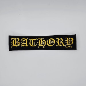 May include: Black rectangular patch with the word "BATHORY" embroidered in gold gothic lettering. The patch is likely for clothing or accessories.