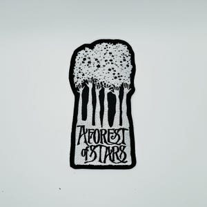 May include: A black and white embroidered patch featuring a stylised forest of trees with star-shaped leaves. The text A FOREST OF STARS is stitched in a decorative font at the base of the tree trunks, all enclosed within a black border.