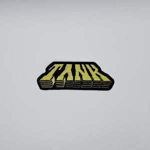 Könnte beinhalten: Gestickter Aufnäher mit dem Wort "TANK" in gelben, gestapelten Buchstaben. Der Aufnäher hat einen schwarzen Rand und ist vor einem weißen Hintergrund platziert. Die Buchstaben haben einen 3D-Effekt.