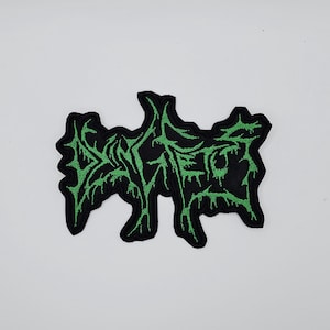 Puede incluir: Parche bordado negro con un diseño verde estilizado. El parche tiene una forma irregular y presenta un patrón orgánico detallado en hilo verde brillante sobre un fondo negro. El diseño parece ser una palabra o símbolo estilizado.