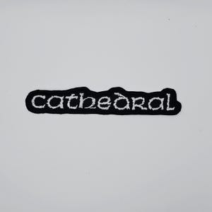 May include: A black patch with white embroidered text reading "Cathedral". The patch has a black border and a rectangular shape. The text is in a decorative, stylized font.