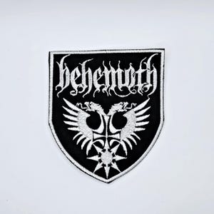 May include: Black and white embroidered patch with the word "Behemoth" in ornate lettering. The patch features a shield shape with a symmetrical design of two winged creatures and a starburst.