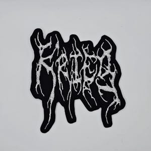 May include: Black embroidered patch featuring white, stylized text. The patch has an abstract design with a dark background and detailed white stitching. Suitable for clothing or accessories.