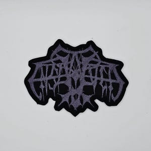May include: A black patch with a stylized, gothic-style design in shades of gray. The design features intricate, interwoven lines and shapes, with the word "ANTAGONISM" in a stylized font. The patch has a black border.