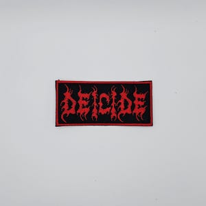 May include: Black rectangular patch with the word "DEICIDE" embroidered in red, stylized, gothic lettering. The patch has a black border and is set against a white background. The lettering has a flame-like design.