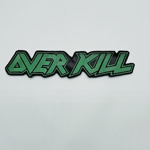 May include: An embroidered patch featuring the word OVERKILL in a stylised, angular font. The lettering is bright green with a thick black border, creating a sharp contrast. The design is elongated and horizontal, with distinct, jagged edges.