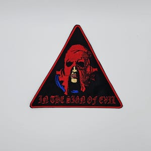May include: Black triangular patch with a red border. The patch features a red, stylized skull design with blue and gold accents. The text "IN THE SIGN OF EVIL" is embroidered in red.