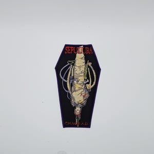 May include: A black, coffin-shaped patch with the band name "SEPULTURA" in red, curved text. The patch features a detailed illustration of a figure with tubes and bandages, with the text "CHAOS A.D." beneath the figure.