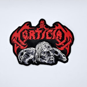 Puede incluir: Parche bordado con la palabra "MORTICIA" en rojo, letras estilizadas con un diseño de calavera y alas de murciélago. Debajo, una imagen en blanco y negro de una figura de zombi.