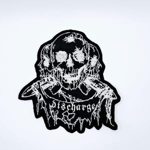 Op de afbeelding: Zwart-wit geborduurde patch met een schedelontwerp en het woord "discharge" in gotisch lettertype. De patch heeft een gekartelde rand en is bedoeld voor kleding of accessoires.
