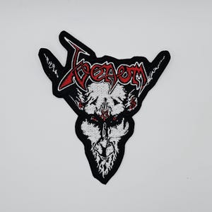 Op de afbeelding: Geborduurde patch met het woord "Venom" in rode, gestileerde letters boven een wit, demonisch gezicht met rode accenten. De patch heeft een zwarte rand en een unieke, hoekige vorm, geschikt voor jassen of tassen.